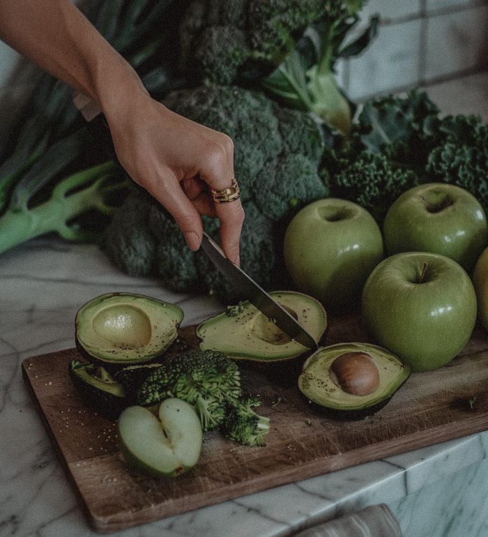 A photo of a woman's hands with gold rings, using one large knife to cut an avocado on a wooden board surrounded by green apples and broccoli, against a white marble kitchen counter. The background features a pile of fresh vegetables including avocados, broccoli, kale leaves, in the style of different artists. --ar 59:74 Job ID: 08af173f-2471-4ea4-94a8-82d8a7905250