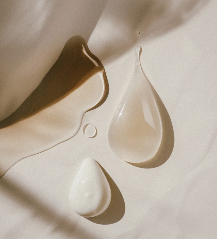 A flat lay of two droplets of cream and oil on an off-white background, with gentle shadows cast in the style of the light source, creating a minimalist aesthetic. The focus is on texture and color contrast between these three elements. Job ID: eea1e021-12d0-4c03-94d2-89b44ae414b0